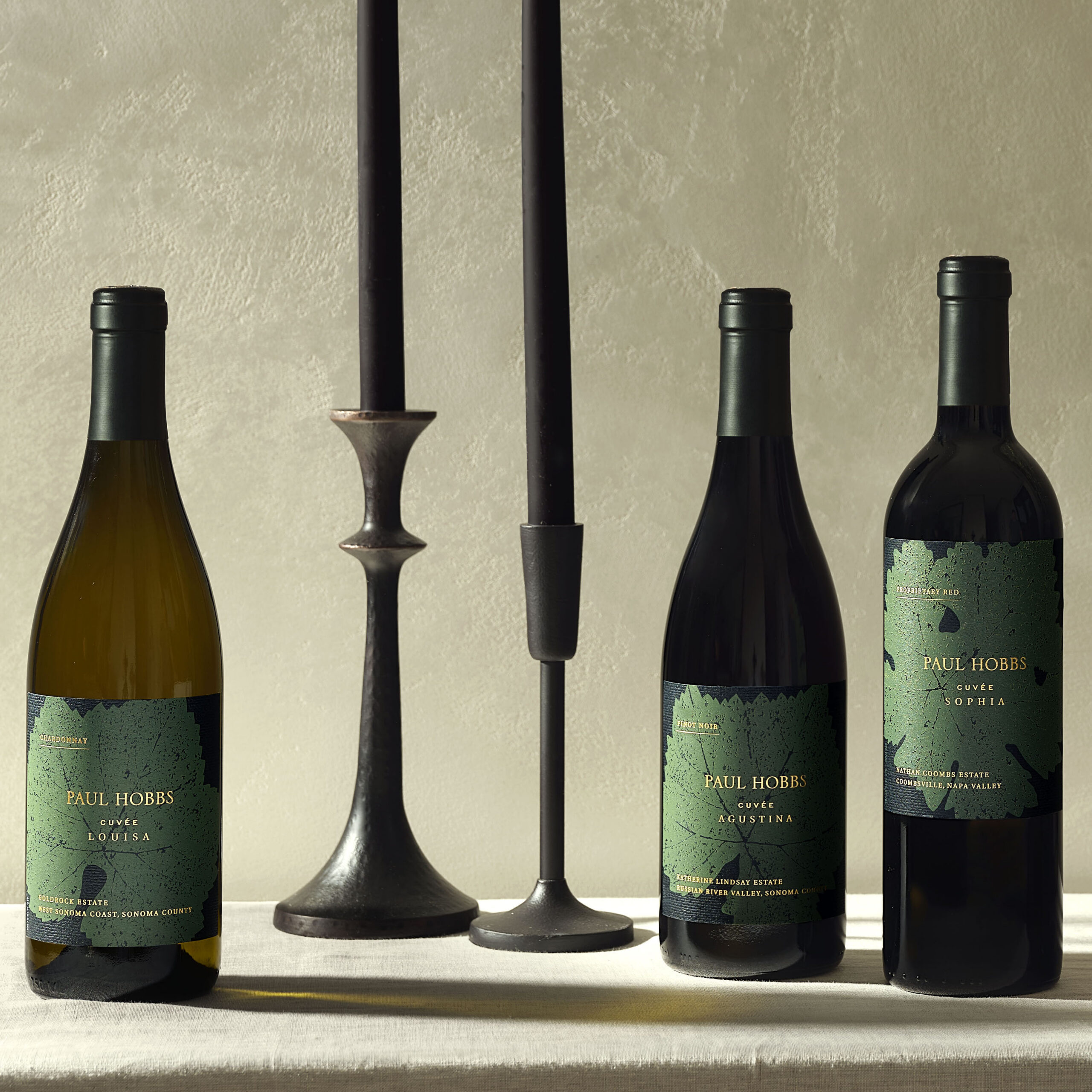 three wine bottles on table with tan linen, with two tall black candlesticks