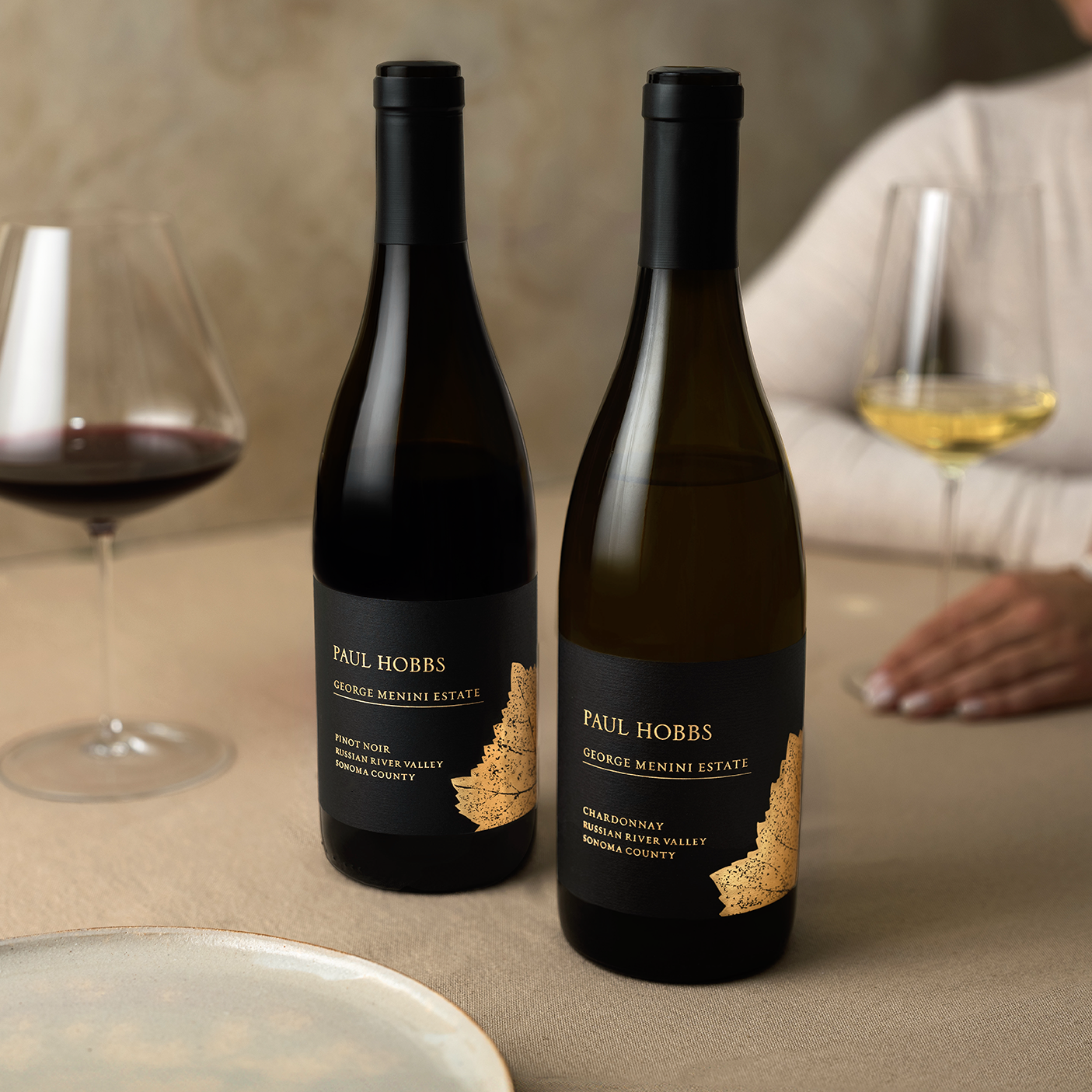 two wine bottles on table with tan linen, a plate and two glasses with wine.
